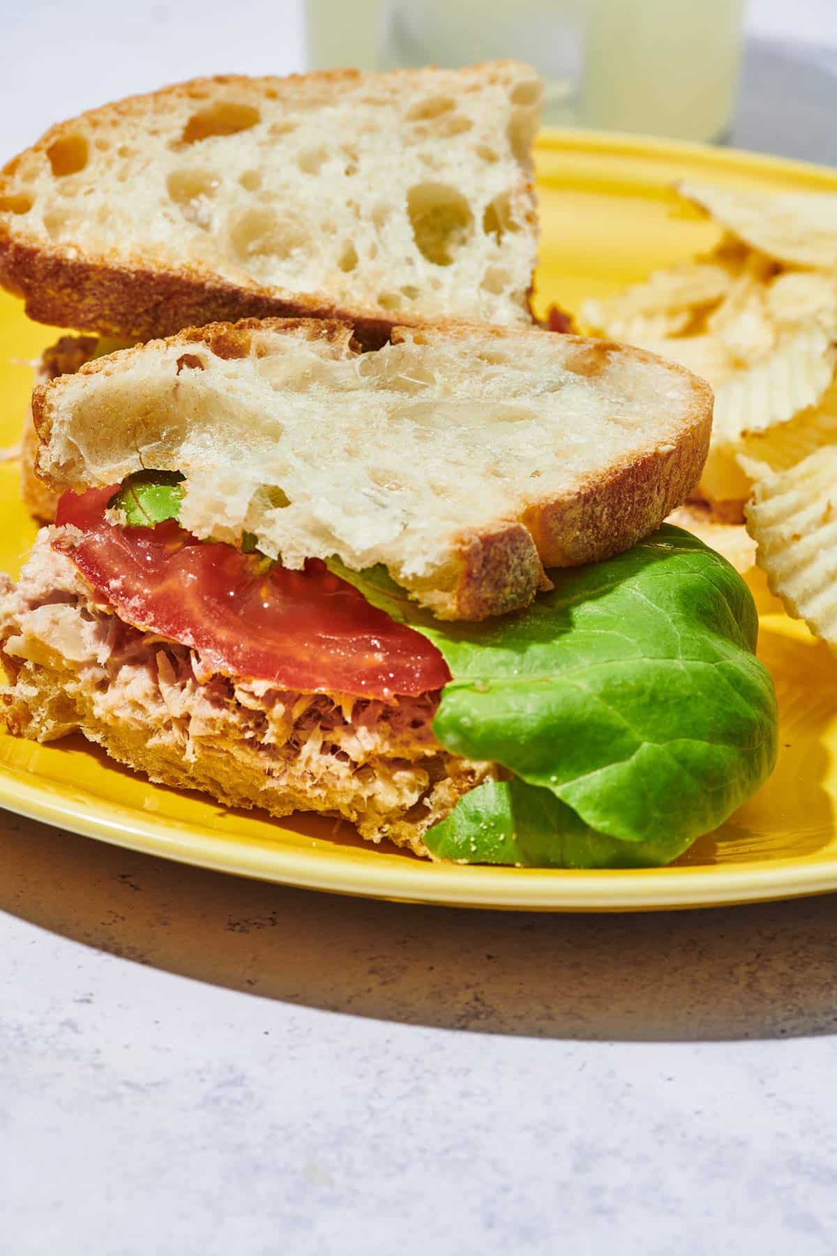 Tuna fish sandwich with tomato and lettuce on yellow plate with chips.