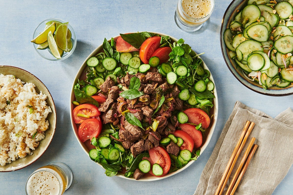 Table setting featuring plate of stir-fried shaking beef with vegetables, bowls of rice and cucumbers, lime wedges, and beer.