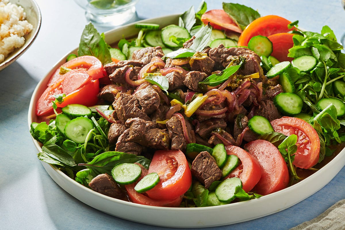 Stir-fried Vietnamese shaking beef on plate over bed of greens, tomatoes, and cucumbers.
