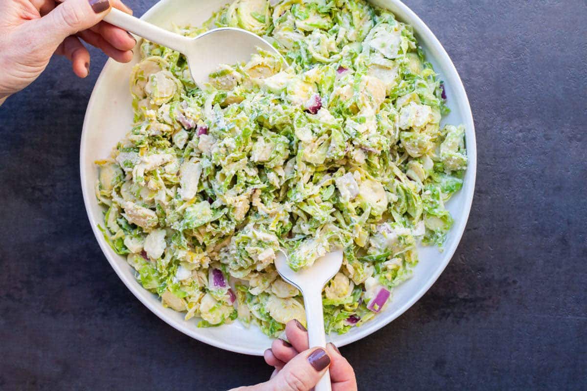 Woman tossing shredded Brussels sprouts with creamy dressing in bowl using spoons.