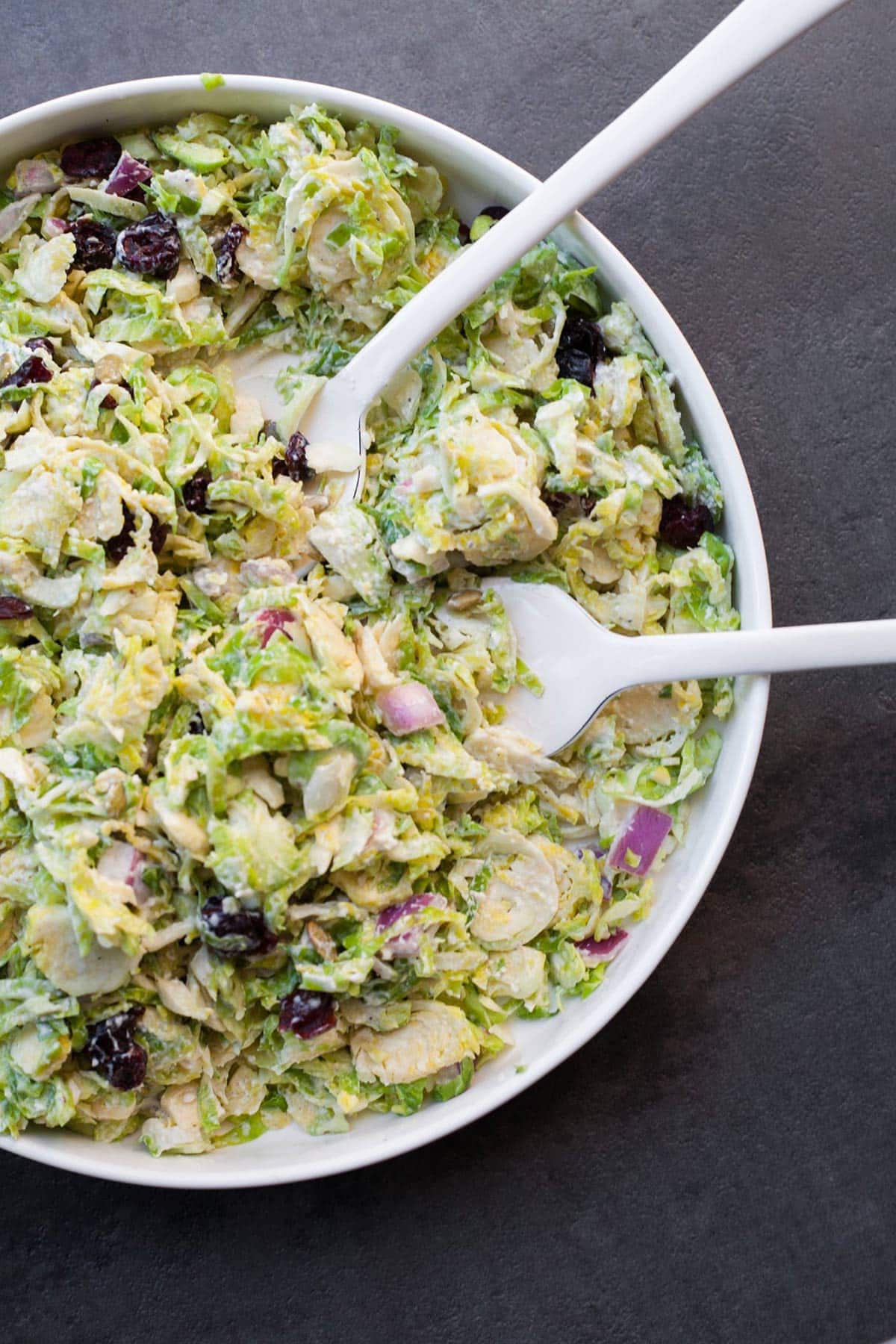 Brussels sprouts slaw in white bowl with white serving spoons.