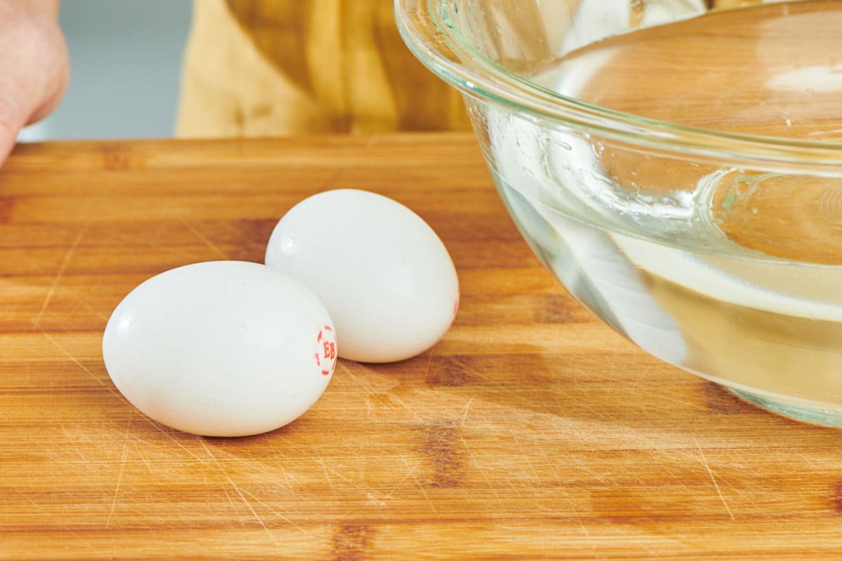 Two eggs on cutting board next to bowl of water.