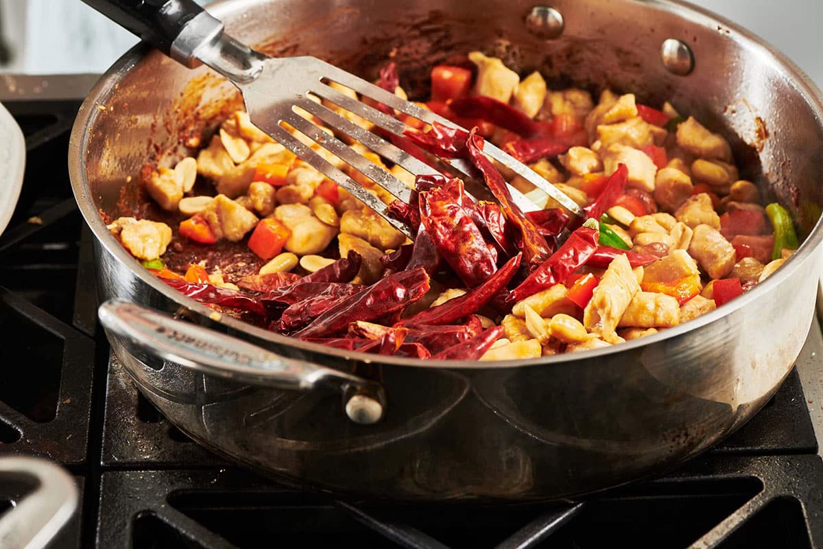 Stirring dried red chili peppers into chicken and vegetables with metal spatula.