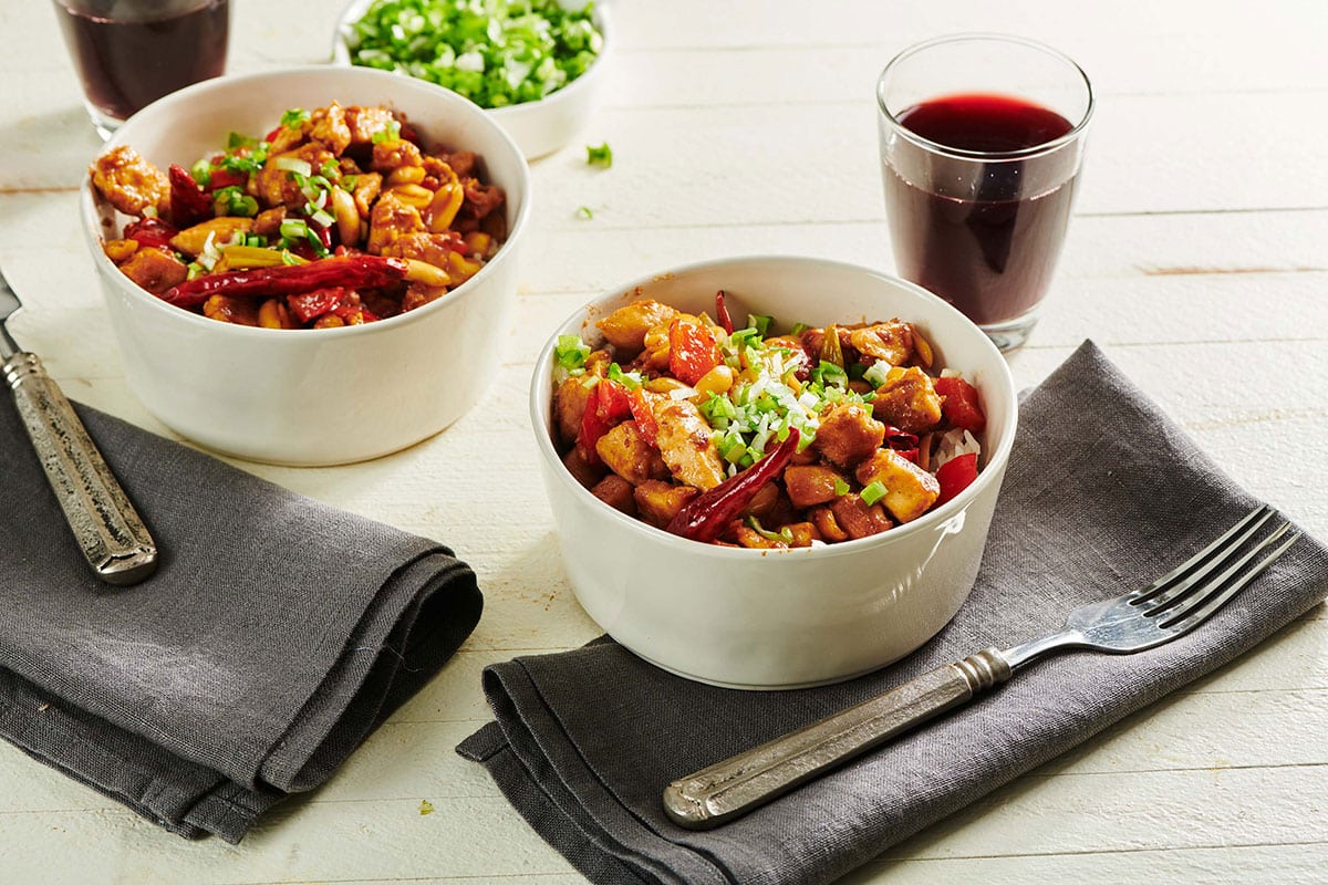 Two bowls of Kung Pao Chicken on table with napkins, forks, and wine.