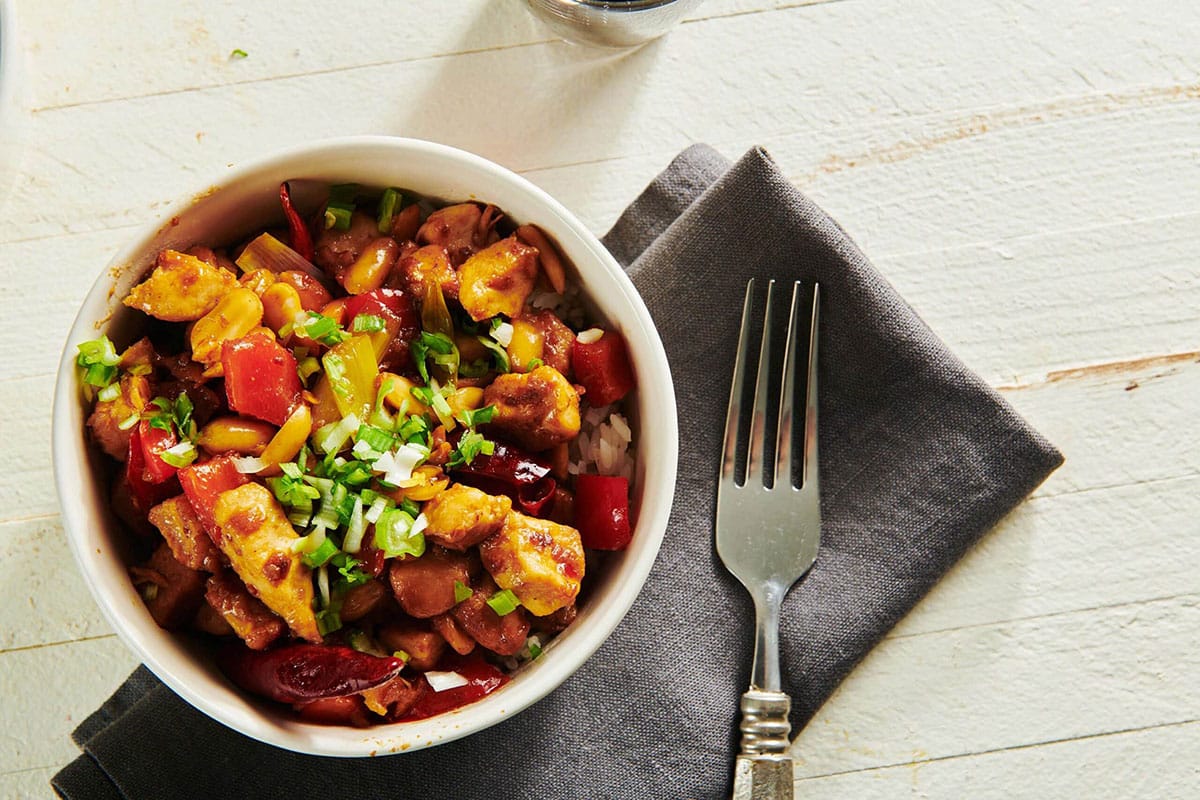 Homemade Kung Pao Chicken in white bowl with grey napkin and fork on table.