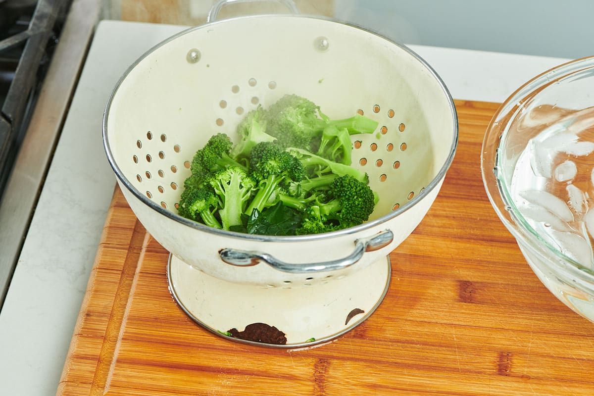 Steamed broccoli in colander on cutting board.