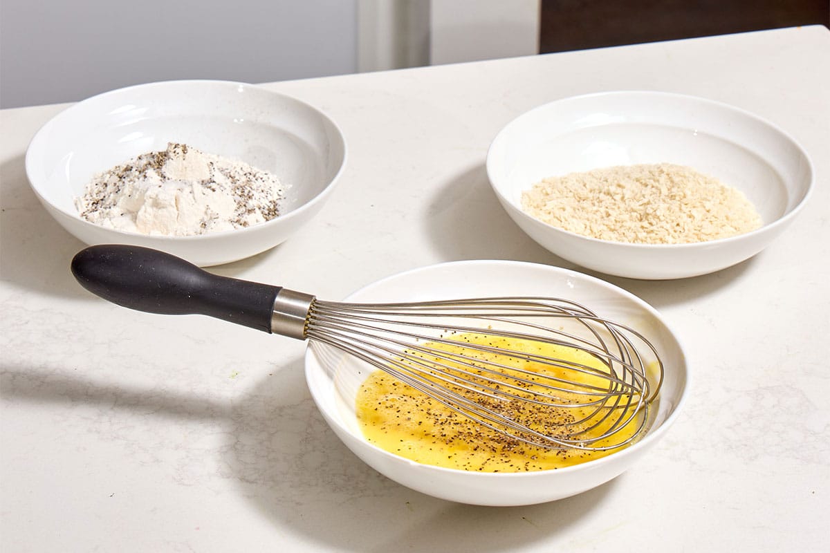 Seasoned flour, beaten eggs, and breadcrumbs in shallow white bowls on counter.