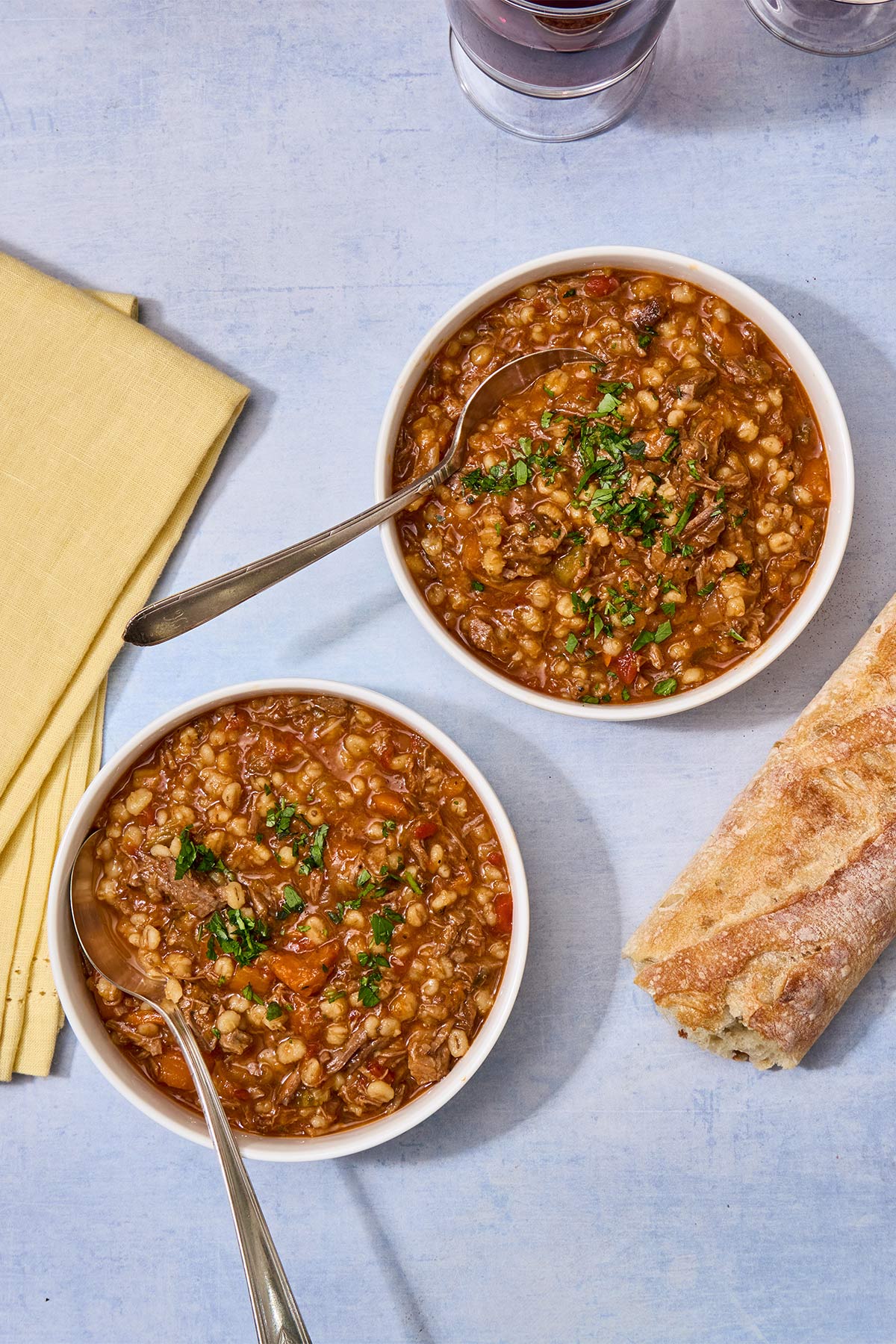 Two bowls with brisket barley soup on table with bread and napkins.