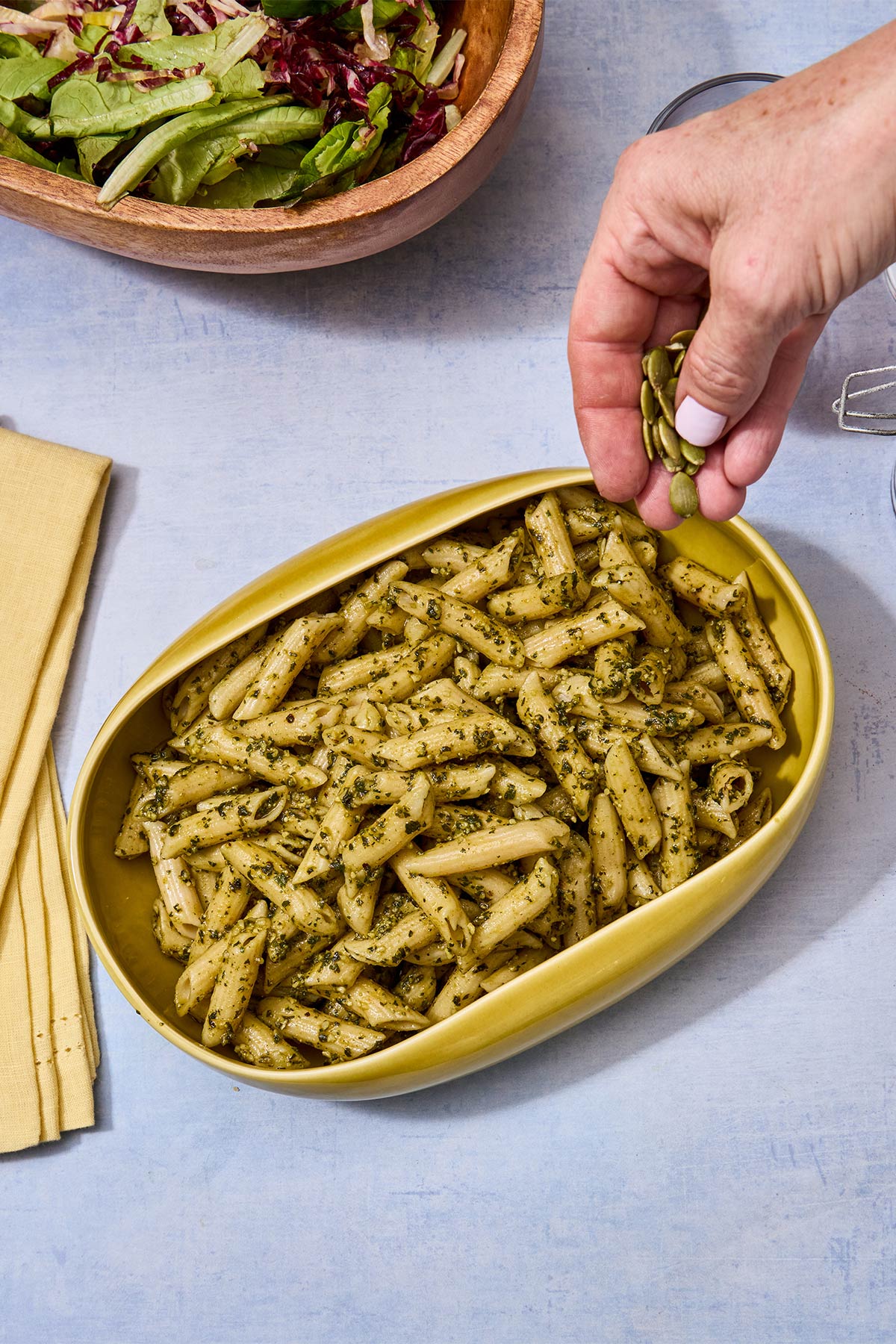 Woman sprinkling pepitas over penne with pumpkin seed pesto in yellow dish.