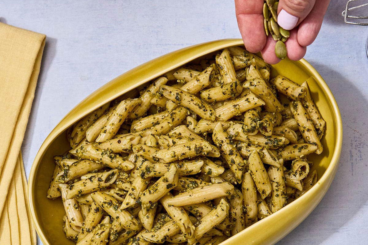 Woman sprinkling pepitas over penne with pumpkin seed pesto in yellow dish.
