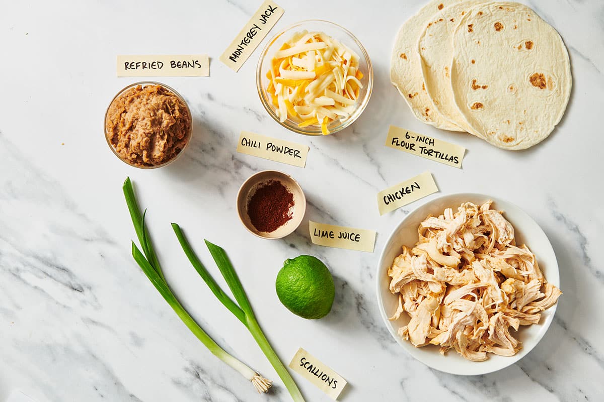 Tortillas, shredded chicken, cheese, lime, and other flauta ingredients on table.