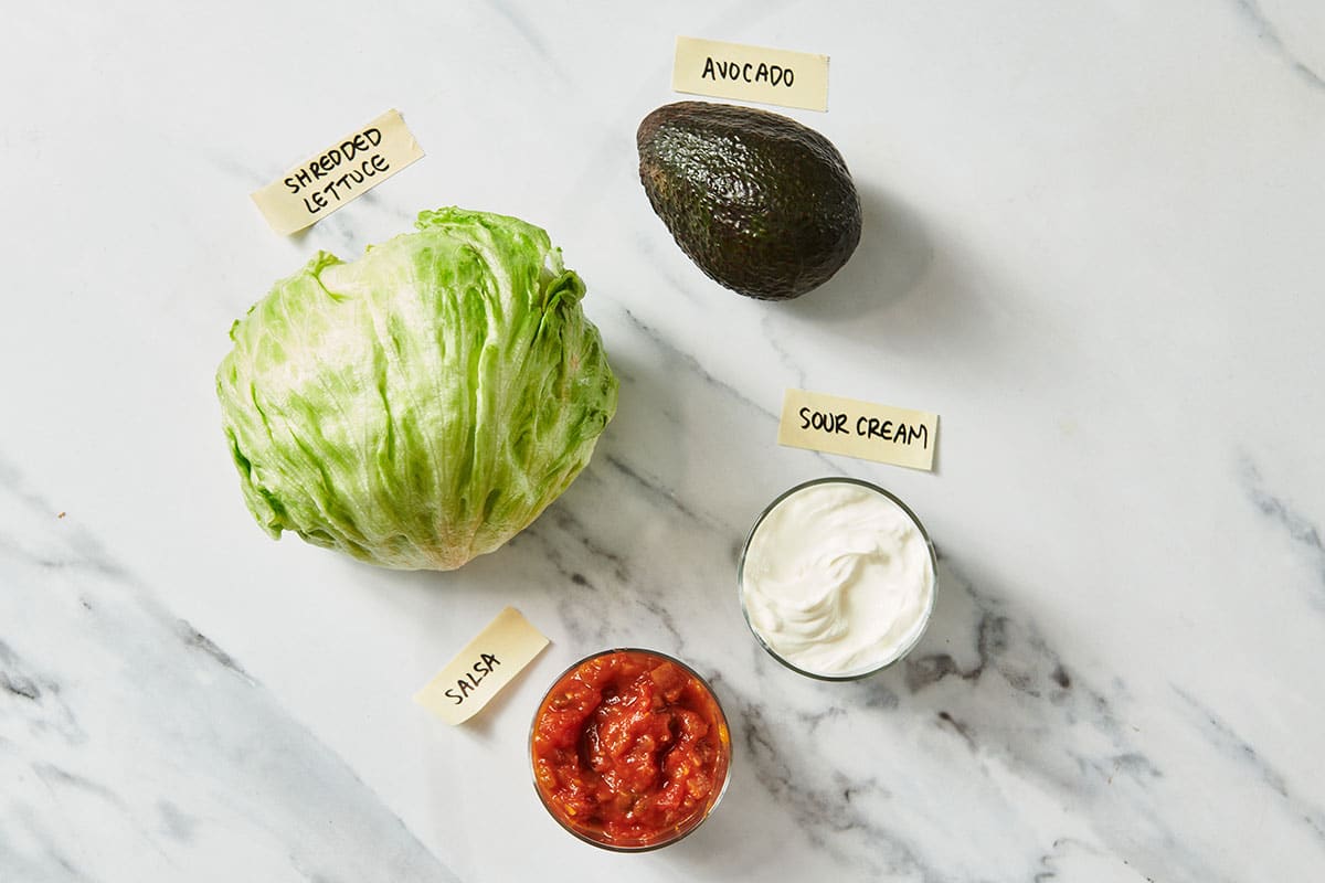 Lettuce, avocado, salsa, and sour cream on white table.