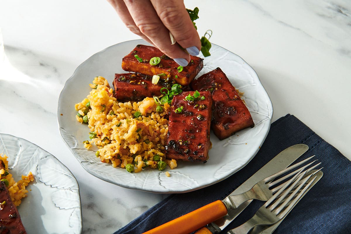Sprinkling Chinese BBQ air-fried tofu with chopped scallions on plate next to rice.