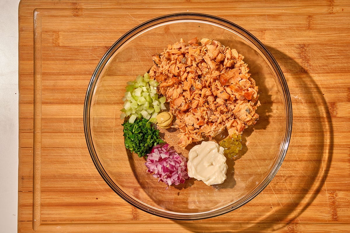 Salmon, mayo, onion, celery, and other salmon salad ingredients in glass bowl.