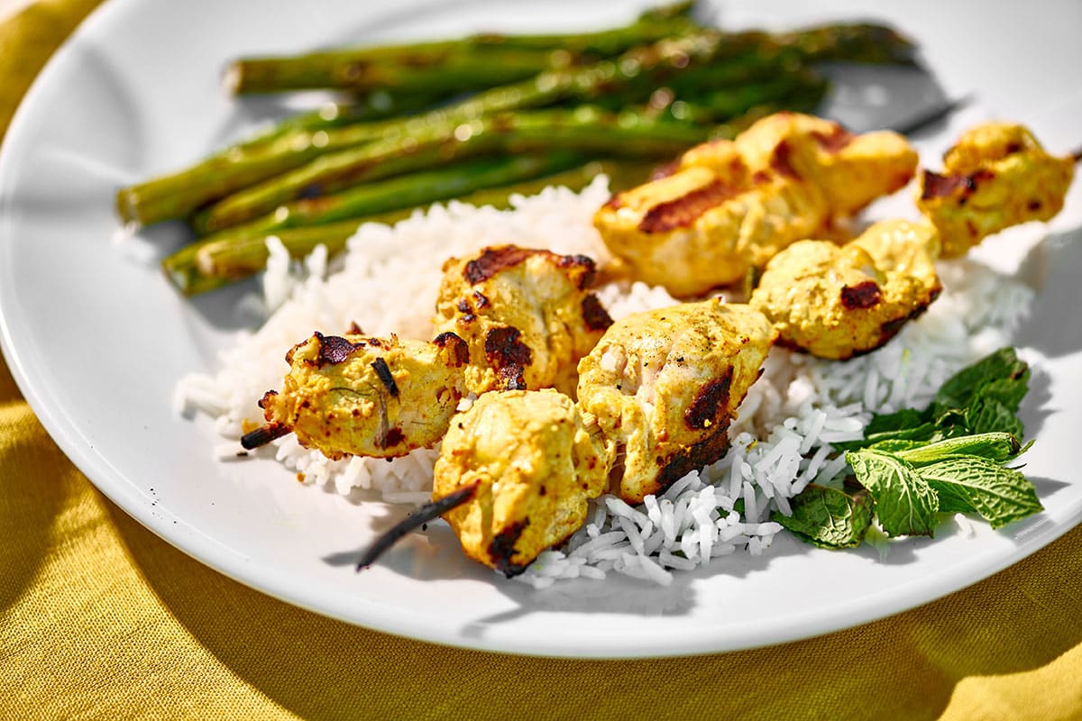 Grilled tandoori chicken skewered on plate with asparagus and other sides.
