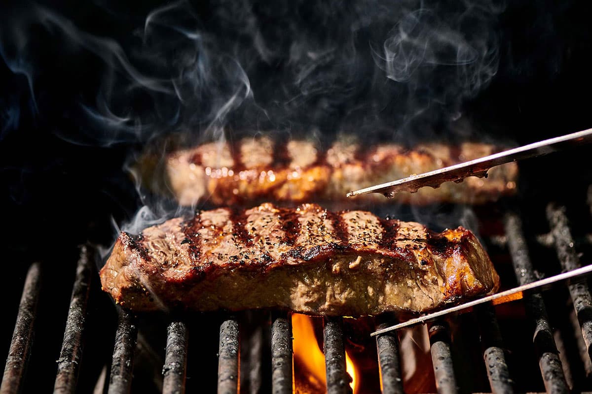 Cooking NY strip steaks on grill and flipping with tongs.