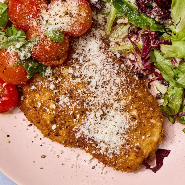 Breaded chicken cutlet on pink plate with salad and tomatoes.