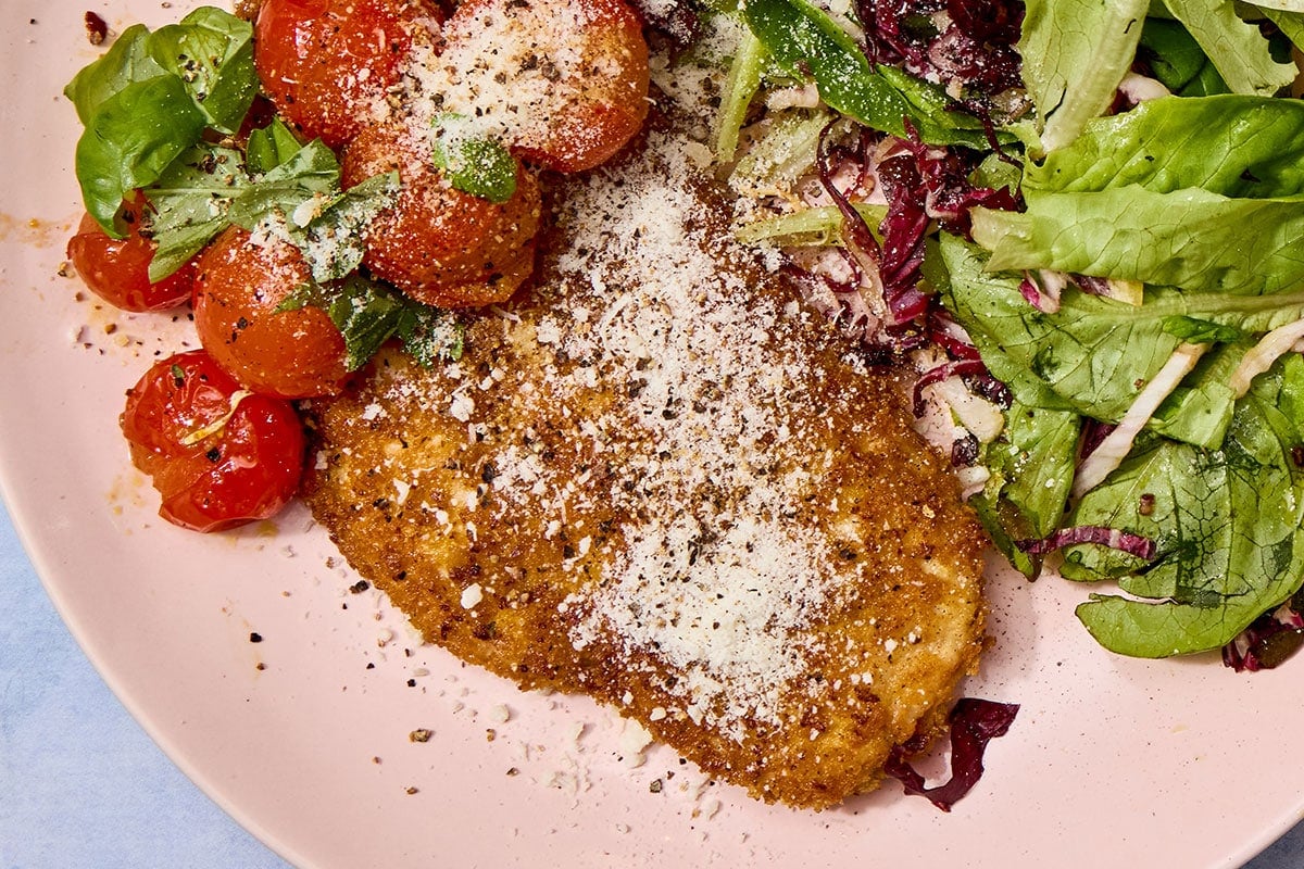 Breaded chicken cutlet on pink plate with salad and tomatoes.