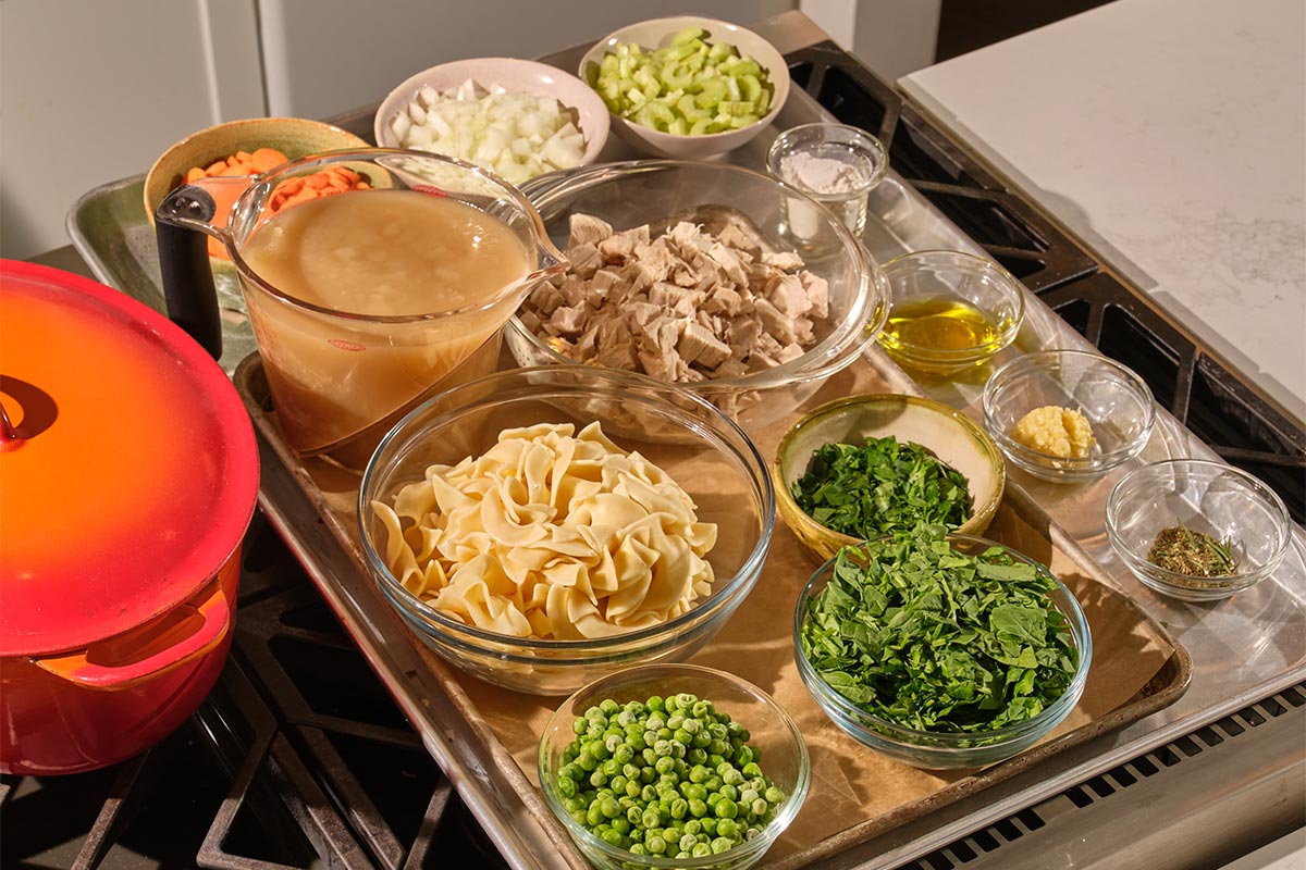 Cooked turkey, broth, peas, fresh herbs, noodles, and other soup ingredients on tray.