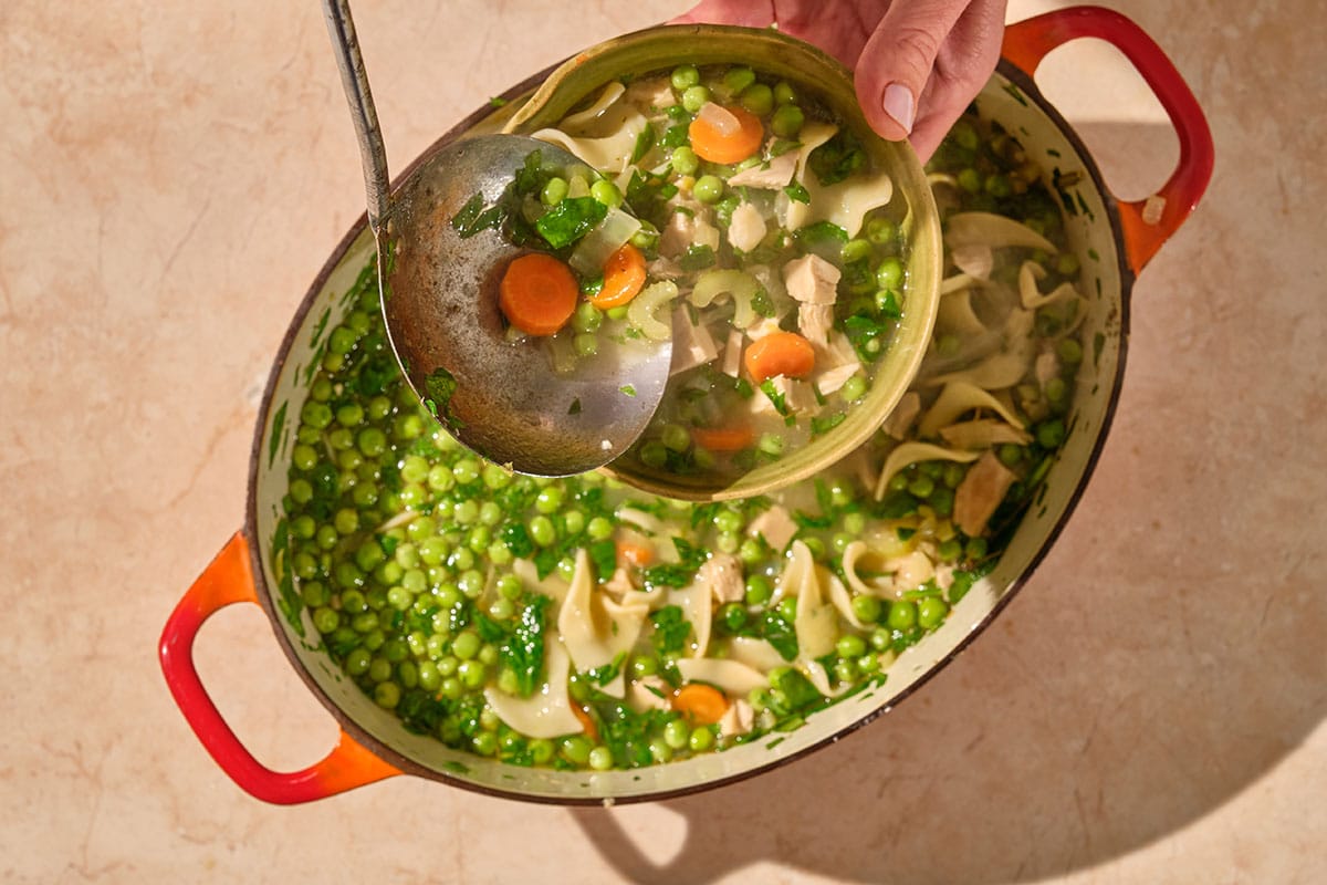 Woman serving turkey noodle soup from dish to bowl with ladel.