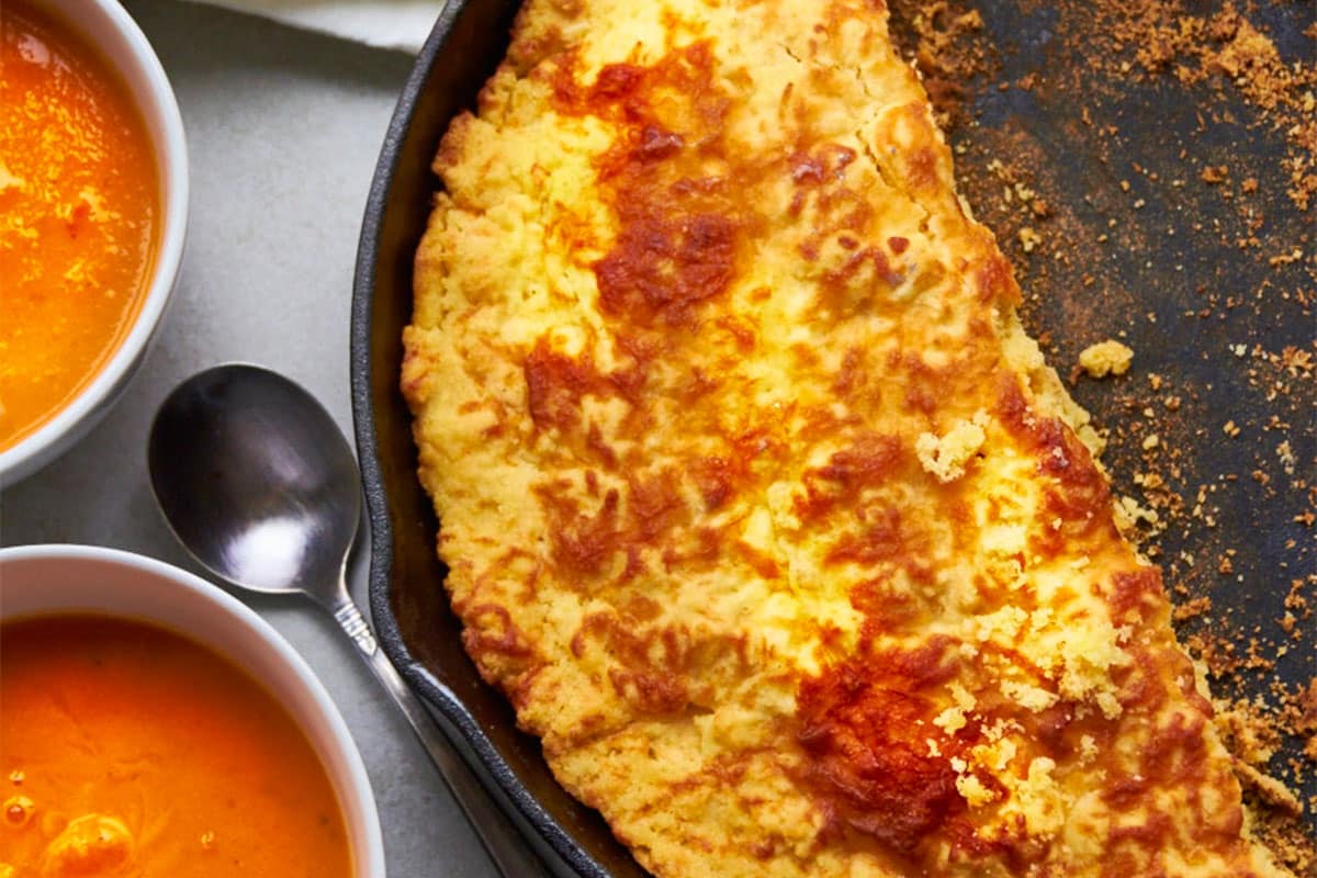 Cheddar cornbread in cast iron skillet next to bowls of tomato soup.