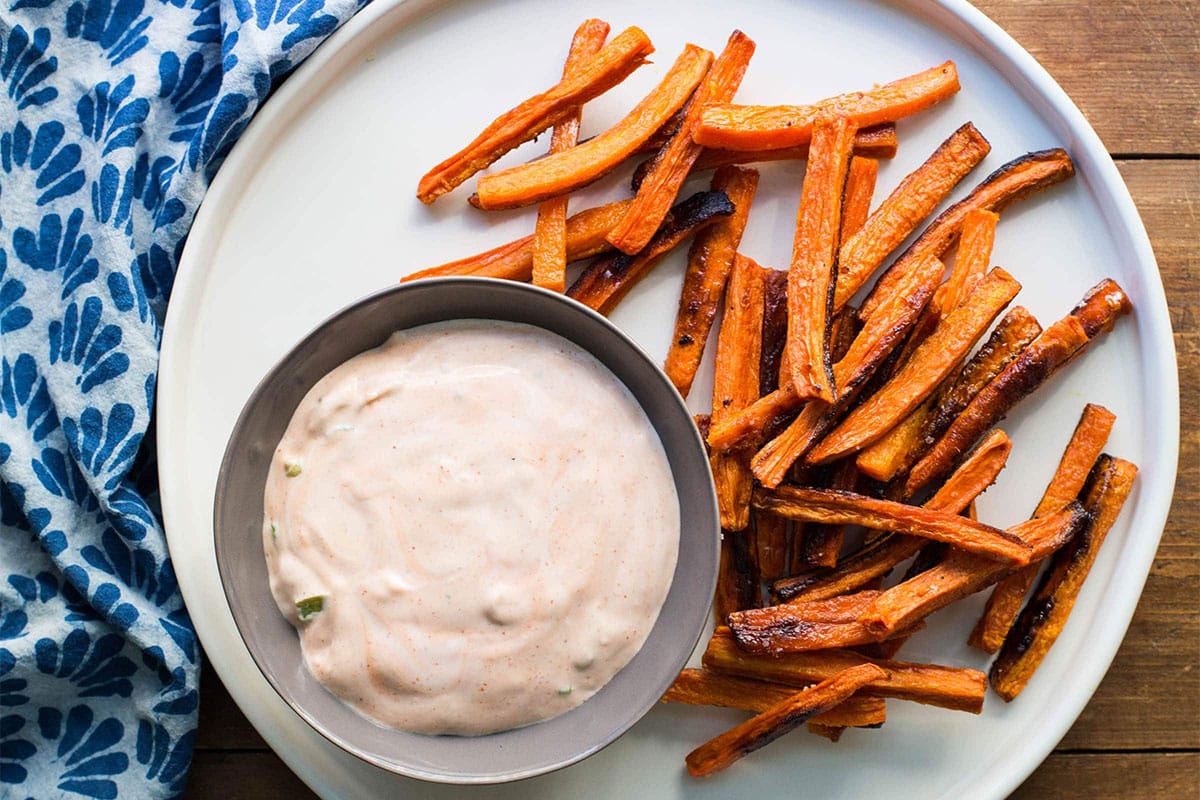 White plate with carrot fries and Sriracha sauce on table with blue napkin.