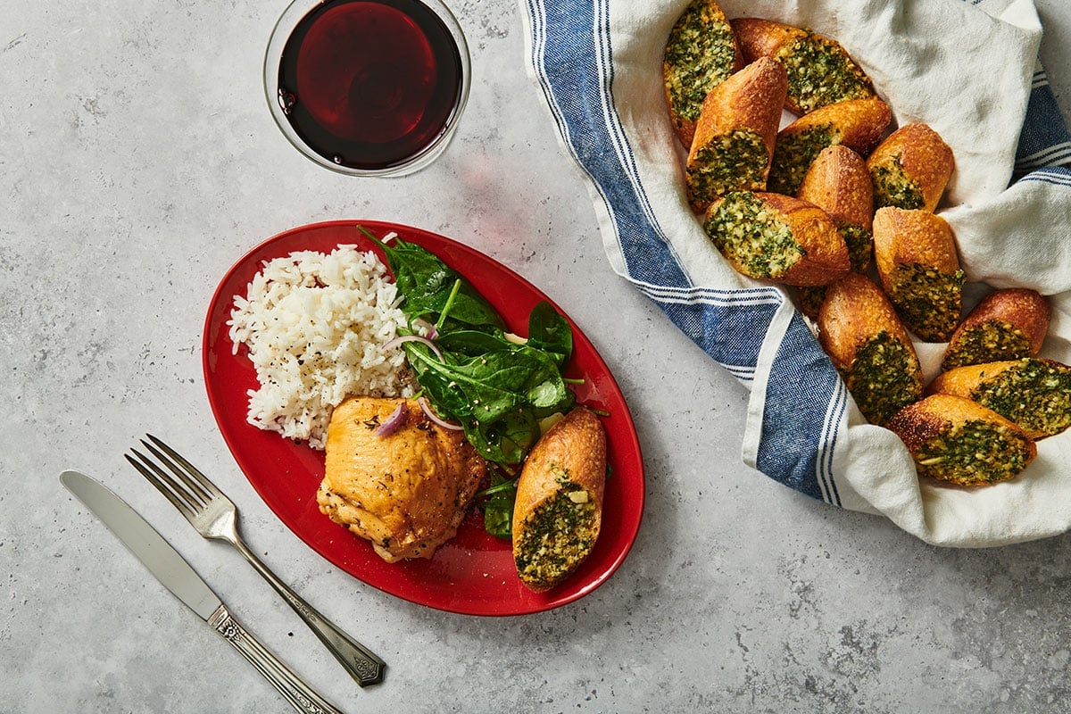 Garlic bread slice on red plate with chicken, salad, and rice next to basket of garlic bread and wine glass.