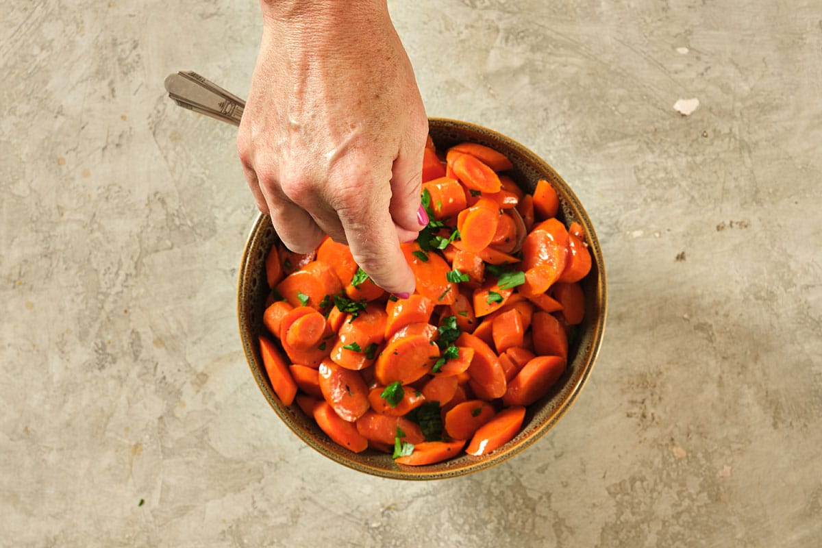 Woman adding fresh chopped herbs to bowl filled with glazed carrots.