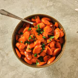 Maple-glazed carrots in bowl with spoon on tan table.