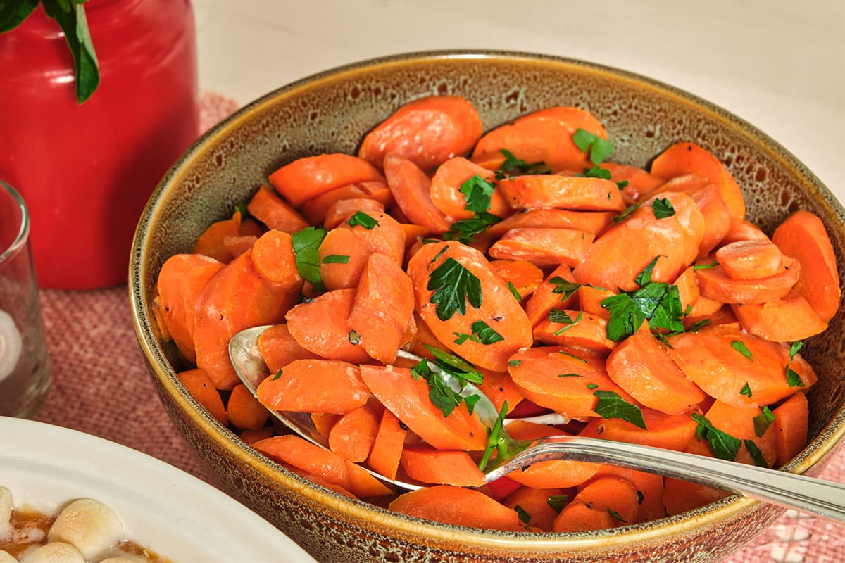 Glazed carrots and herbs in brown bowl with spoon on table.