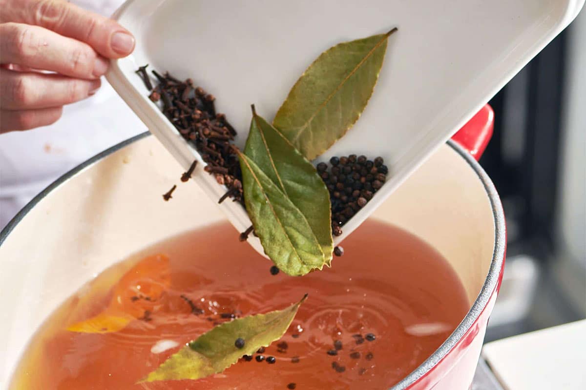 Pouring bay leaves and other herbs and spices into pot of homemade broth.