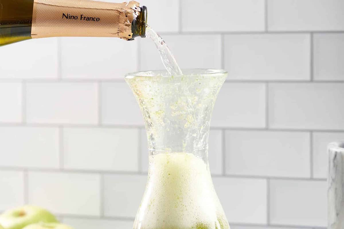 Bottle pouring Prosecco into a glass pitcher of green liquid.