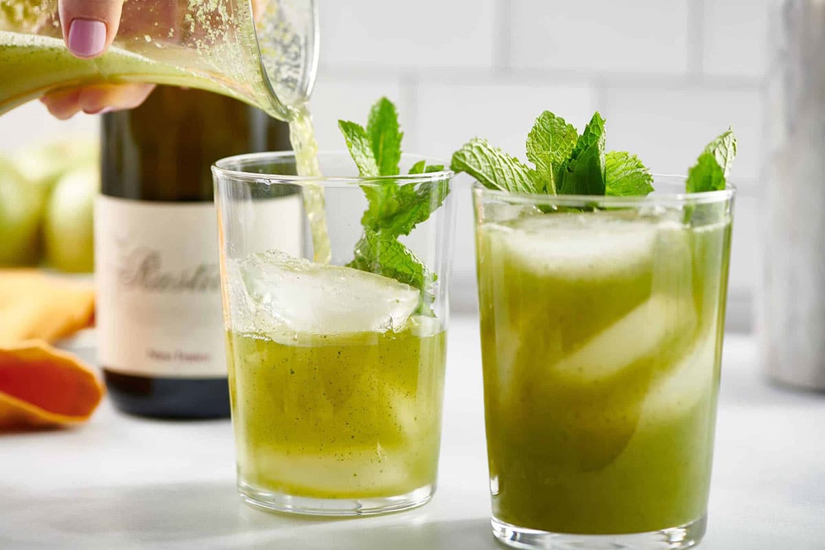 Glass pitcher pouring green Vodka Prosecco cocktails into small glasses of ice.