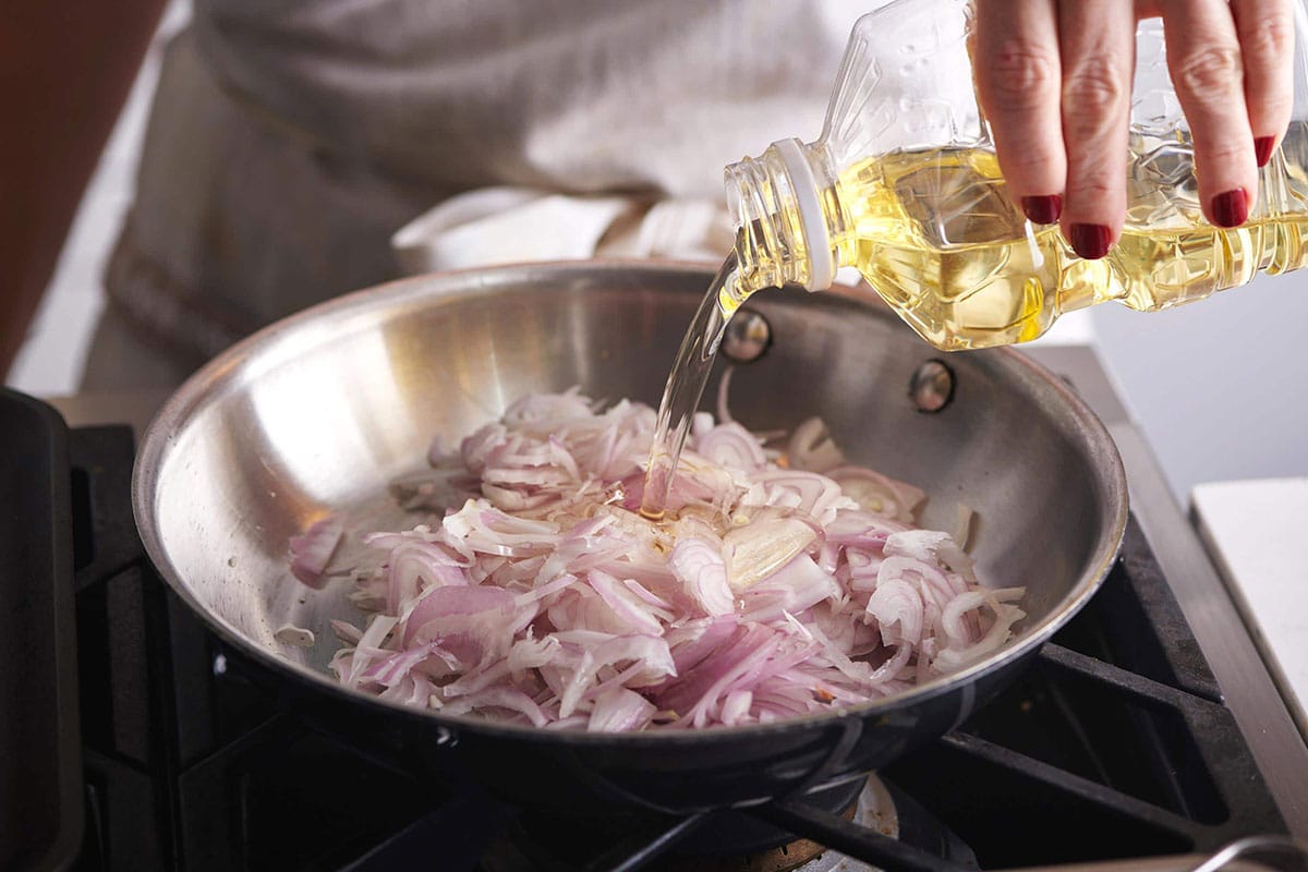 Pouring oil over sliced shallots in pan on stove.
