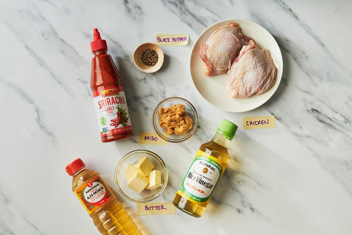 Chicken thighs, miso, hot sauce, and other ingredients on white marble table.