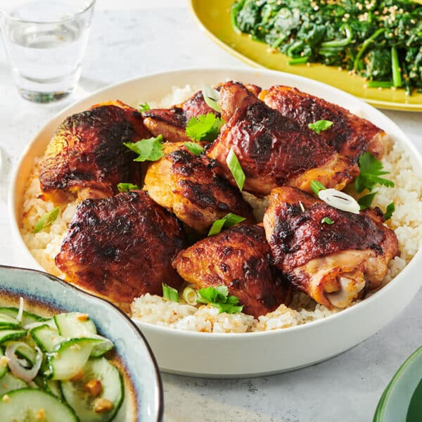 Miso chicken thighs on white serving dish over rice with Asian salads on table.
