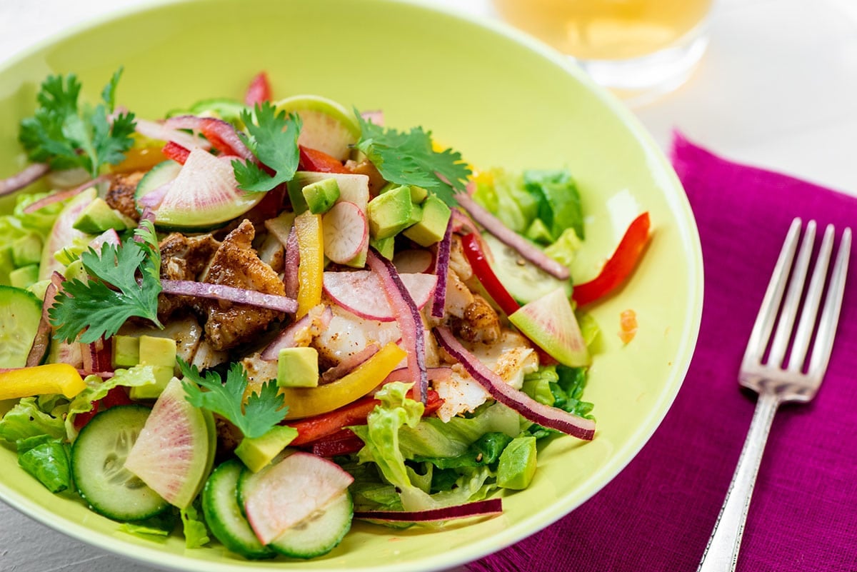Yellow bowl filled with a fish taco salad on table with pink napkin and fork.