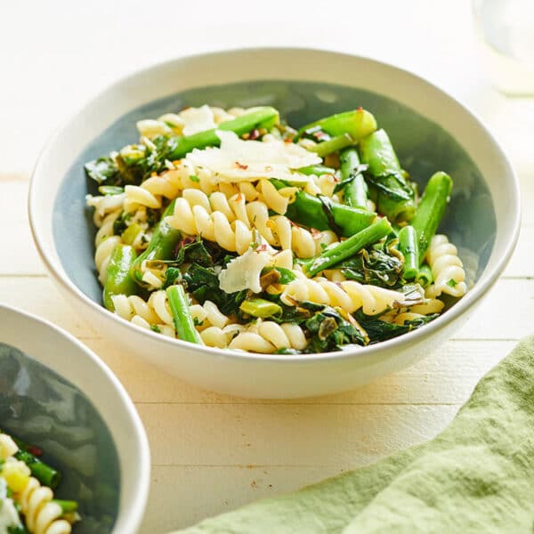 White bowls on table filled with rotini pasta, green beans, snap peas, and Swiss chard in goat cheese sauce.
