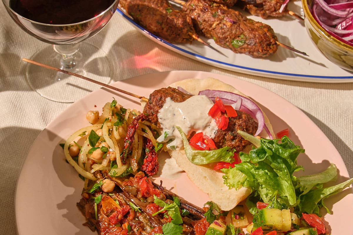 Garlic yogurt sauce atop lamb kofta on pink plate with Turkish side dishes.