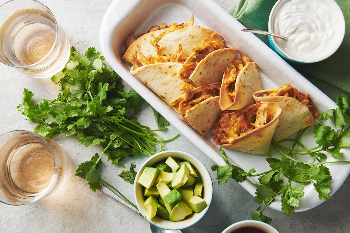 Baked chicken quesadillas in dish on table with toppings and drinks.