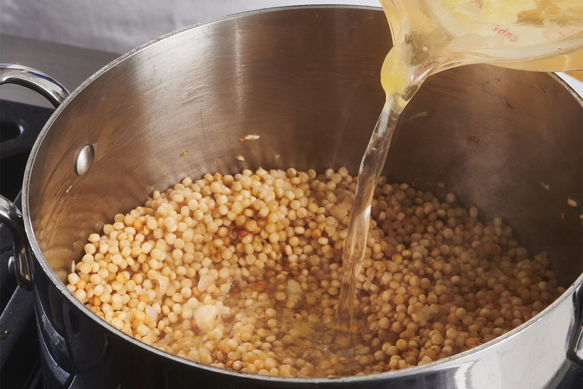 Pouring broth into a pan of Israeli couscous cooking in pot on the stove.
